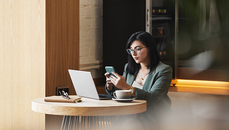 a lady sitting on her phone at a cafe with her laptop and a coffee in front of her